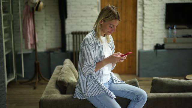 Blonde-Haare-kaukasische-Frau-sitzt-auf-dem-Sofa-im-Wohnzimmer-mit-Smartphone-für-das-Senden-von-Nachricht-im-Online-Chat