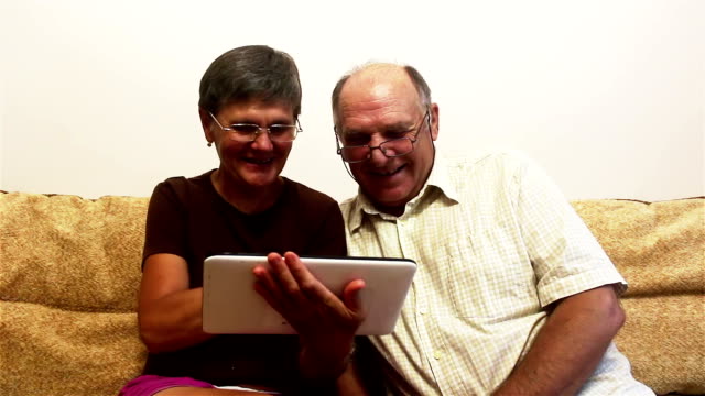 An-elderly-couple-is-sitting-on-a-sofa-at-home-and-watching-photos-on-a-tablet-pc,-smiling-and-talking-actively.