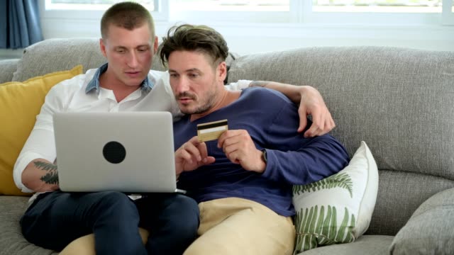 Pareja-gay-relajándose-en-el-sofá-usando-la-computadora-portátil.-Trate-de-completar-la-compra.