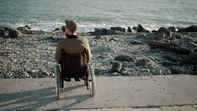 Alone-male-wheelchair-user-is-sitting-calmly-on-seacoast-and-relaxing,-back-view