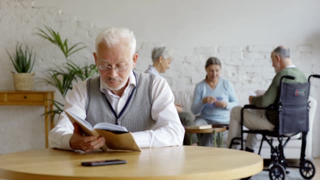 Rack-Fokus-von-drei-älteren-Menschen,-zwei-Seniorinnen-und-behinderten-alten-Mann-im-Rollstuhl,-Spielkarten-in-betreutem-Wohnen.-Intelligenter-alter-Mann-in-Brille-lesen-Buch-und-denken-am-Tisch