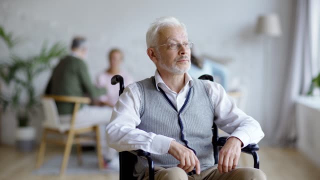 Tracking-medium-shot-of-disabled-senior-man-in-eyeglasses-sitting-in-wheelchair-looking-away-and-thinking-in-nursing-home,-other-aged-patients-in-background