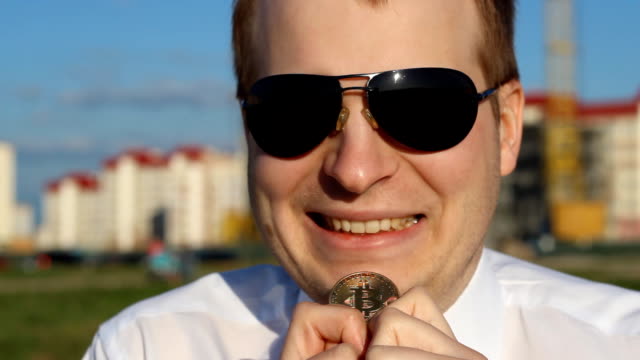 Ein-Mann-in-schwarzer-Brille-hält-einen-Bitcoin-in-den-Händen-und-lacht,-glücklich,-Nahaufnahme