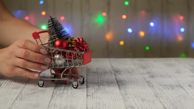 Hand-puts-mini-shopping-cart-with-christmas-tree-and-gifts-on-the-background-of-led-lamps-garland.
