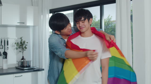 Asian-Gay-couple-standing-and-hugging-room-at-home.-Young-handsome-LGBTQ+-men-kissing-happy-relax-rest-together-spend-romantic-time-in-modern-kitchen-with-rainbow-flag-at-house-in-the-morning-concept.