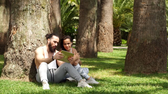 Hombre-y-mujer-relajándose-con-tableta-digital-en-el-parque-después-del-entrenamiento-deportivo