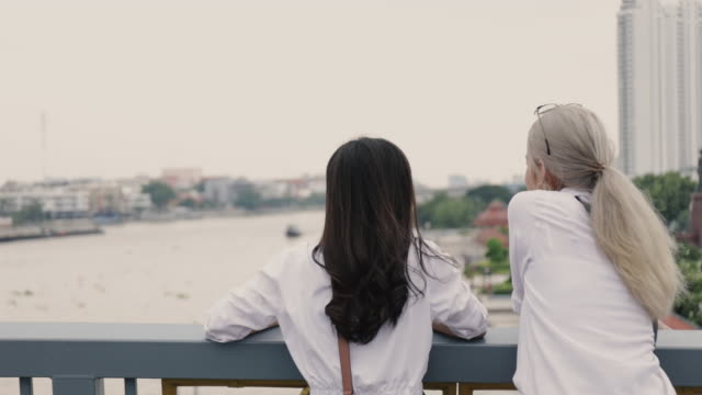 Vista-trasera-de-parejas-lesbianas-asiáticas-disfrutando-de-viajar-y-hablar-mientras-están-de-pie-en-el-puente.-Concepto-LGBT.
