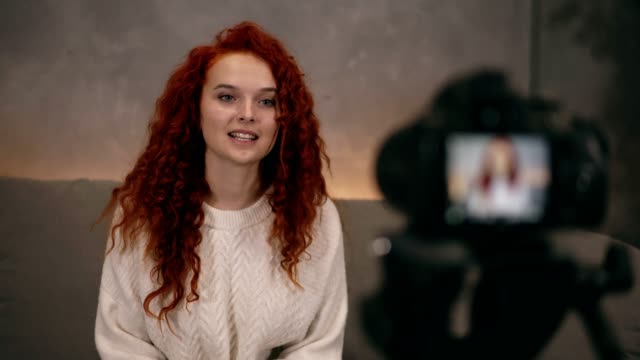 Portrait-of-red-headed-young-girl-vlogger-is-talking-in-front-of-camera-recording-video-for-online-blog-for-her-followers,-smiling-and-gesturing.-Woman-is-wearing-jeans-and-white-sweater.-Accelerated-video.-Camera-on-foreground,-blurred