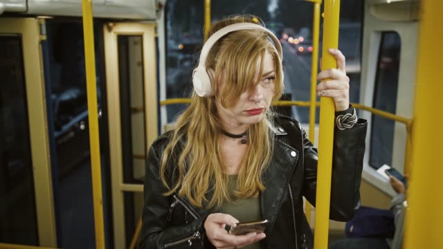 Stylish-sad-woman-hipster-in-headphones-is-listening-to-music.-Riding-tram,-using-cellphone,-standing-and-holding-onto-the-handrail.-Slow-motion