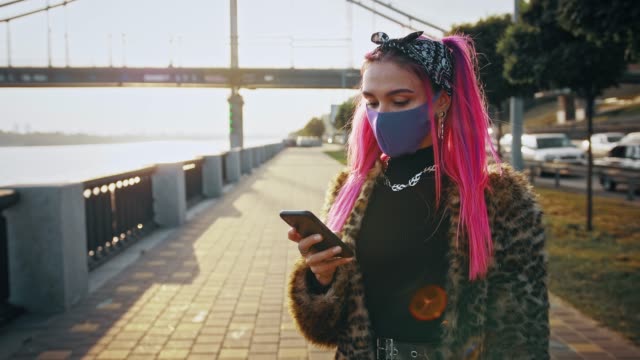 Junge-Hipster-Dame-in-informellem-Outfit-und-Schutzmaske-benutzt-Smartphone,-während-sie-an-der-Böschung-steht