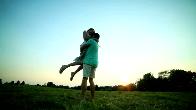 Young-man-spins-and-holds-happy-woman-in-arms-twirling-together-at-sunset-backlit-grass-POV.-Boyfriend-rotates-carrying-smiling-long-hair-girlfriend-in-sunny-evening-summer-meadow.-Love-togetherness