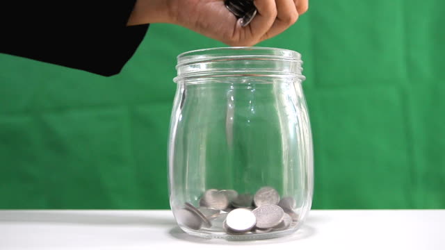 Close-up-shot-hand-of-woman-drop-the-coins-to-glass-jar-over-green-background