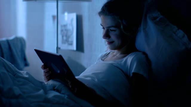 Young-Teenage-Girl-Lies-in-Her-Bed-at-Night-With-Tablet-Computer.-Watches-TV-Show-and-Smiles.