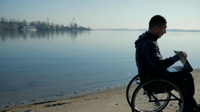 körperlich-Behinderte-Mann-sitzt-in-einem-Rollstuhl-am-Meeresufer,-in-Händen-laptop