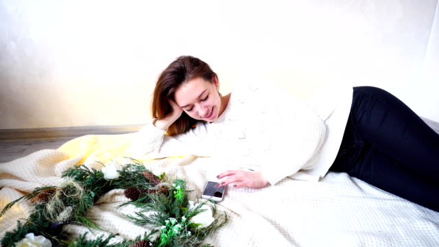 Stunning-female-correspond-in-social-networks-using-smartphone,-lying-on-floor-on-plaid-next-to-Christmas-wreath-in-cozy-room-with-garland