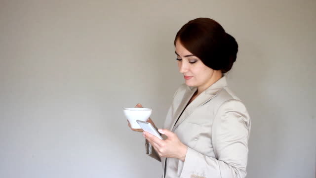 Beautiful-young-businesswoman-looks-at-the-phone-screen-and-plays-downloaded-application-on-a-smart-phone.-Elegant-woman-communicate-in-social-networks-and-drinks-coffee