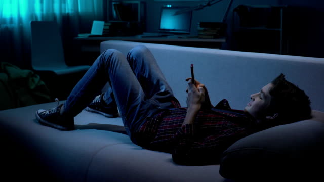 Young-male-lying-on-couch-and-chatting-with-friend-on-smartphone,-social-network