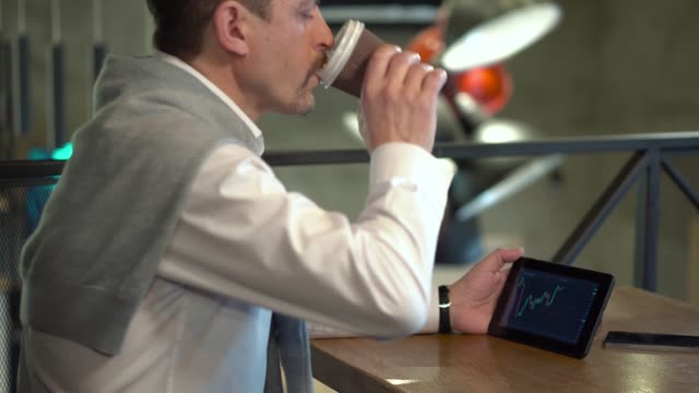 Reife-schöne-Geschäftsmann-überwacht-Handelsplätze-auf-Tablet-und-trinken-Kaffee-in-einem-café