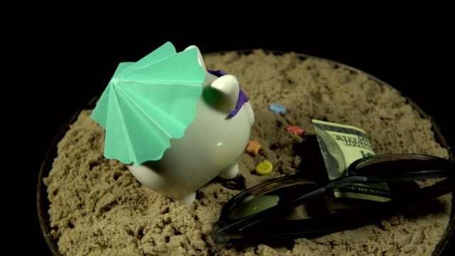A-white-piggy-bank-stands-on-a-sandy-beach-and-revolves-on-a-black-background.