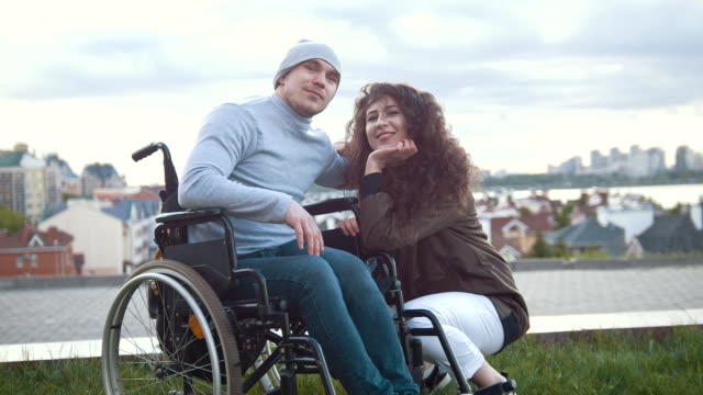 Portrait-of-a-happy-disabled-man-in-a-wheelchair-embraces-with-young-woman-outdoors