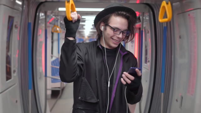 Young-handsome-stylish-man-wearing-black-hat-and-eyeglasses-with-headphones-listening-to-music,-browsing-on-smartphone-in-public-transport,-millenial-hipster-boy-enjoying-mobile-technology-in-subway