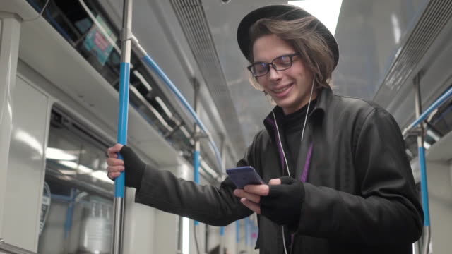 Young-stylish-male-hipster-wearing-black-hat-and-eyeglasses-with-headphones-listening-to-music,-browsing-on-smartphone-in-public-transport,-millenial-boy-enjoying-mobile-technology-in-subway