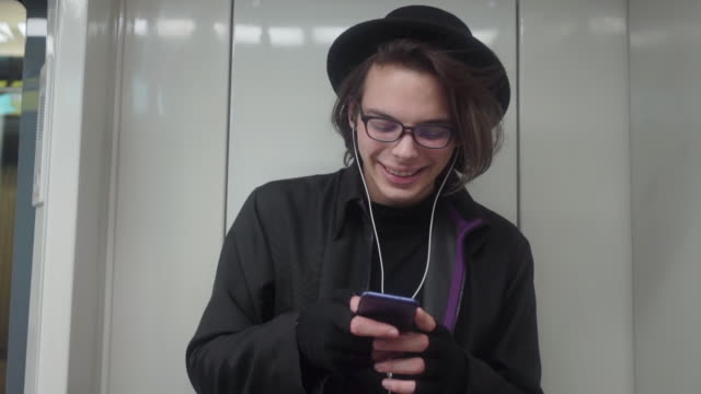 Young-stylish-male-hipster-wearing-black-hat-and-eyeglasses-with-headphones-listening-to-music,-browsing-on-smartphone-in-public-transport,-millenial-boy-enjoying-mobile-technology-in-subway