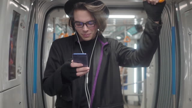 Joven-elegante-hipster-masculino-con-sombrero-negro-y-anteojos-con-auriculares-que-escuchan-música,-navegar-en-el-teléfono-inteligente-en-el-transporte-público,-chico-milenal-disfrutando-de-la-tecnología-móvil-en-el-metro