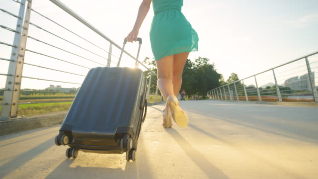 ANGLE-BAJO:-Una-mujer-joven-con-tacones-salta-mientras-camina-hacia-el-aeropuerto-al-atardecer
