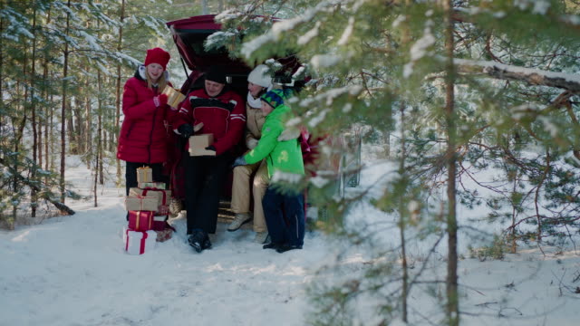 Happy-family-looking-New-Year-gift-in-car-trunk-in-winter-forest.-Mom,-dad,-son-and-daughter-in-red-Christmas-hat-having-fun-together-dog-in-snowy-woodland-at-winter-holiday.