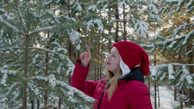 Happy-girl-touching-silver-ball-hanging-on-New-Year-tree-in-winter-forest.-Young-girl-teenager-playing-with-decoration-ball-on-Christmas-tree-in-snowy-forest-at-winter-walk.