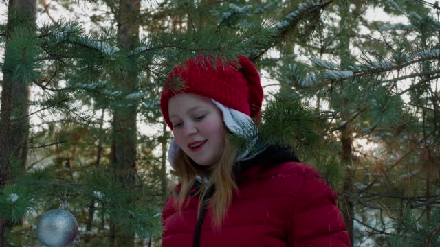 Happy-girl-playing-with-snowy-pine-tree-branch-in-winter-forest.-Young-woman-touching-coniferous-tree-branch-with-Christmas-ball.-Teenager-girl-in-New-Year-woodland.