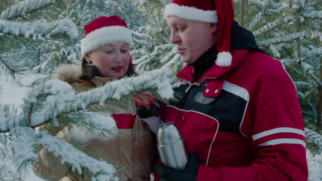 Romantisches-Paar-in-roten-Weihnachtsmütze-trinken-heißen-Tee-aus-Thermoflasche-in-verschneiten-Wald.-Mann-und-Frau-in-der-Liebe-trinken-heißen-Tee-aus-Tasse-im-Nadelwald-im-Winter.