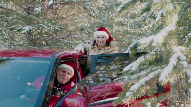 Fröhlicher-Mann-in-rotem-Weihnachtshut-wie-der-Weihnachtsmann-winkt-die-Hand-aus-dem-Auto-im-verschneiten-Wald.-Glückliches-Paar-im-Neujahr-Hut-im-Winterwald-auf-verschneiten-Bäumen-Landschaft.