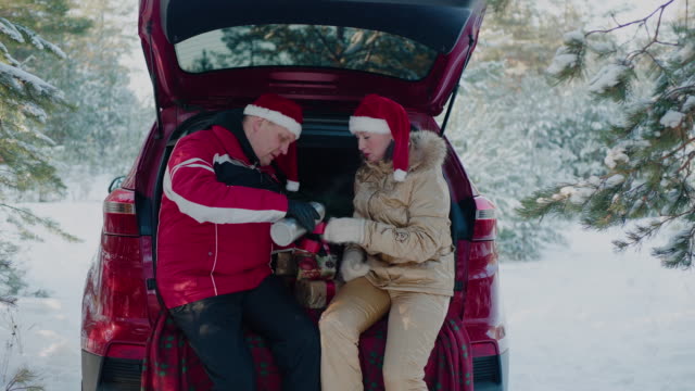 Happy-couple-in-Christmas-hat-drinking-hot-tea-from-thermo-flask-in-car-back-seat-in-winter-forest.-Man-and-woman-in-red-New-Year-hat-drinking-hot-tea-at-showy-woodland.