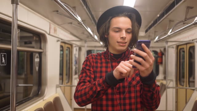 Young-handsome-stylish-man-wearing-black-hat-with-headphones-listening-to-music,-browsing-on-smartphone-in-public-transport,-millenial-hipster-boy-enjoying-mobile-technology-in-subway
