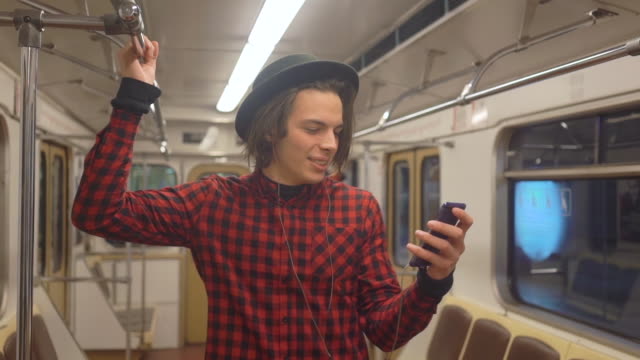 Junge-hübsche-stilvolle-Mann-trägt-schwarzen-Hut-mit-Kopfhörern-Hören-von-Musik,-Surfen-auf-dem-Smartphone-in-öffentlichen-Verkehrsmitteln,-tausendjährige-Hipster-Junge-genießen-mobile-Technologie-in-der-U-Bahn