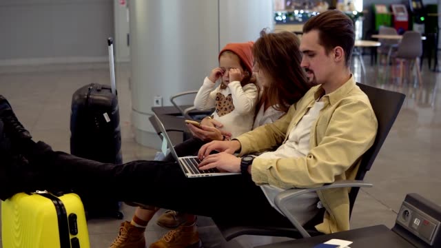 Junger-Vater-arbeitet-auf-Laptop-am-Flughafen-mit-seiner-Familie,-während-auf-die-Abreise-warten.-Langhaarige-Mutter-unterhält-ihre-kleine-Tochter-mit-Smartphone,-so-dass-sie-es-benutzen-kann.-Flughafen-moderne-Halle