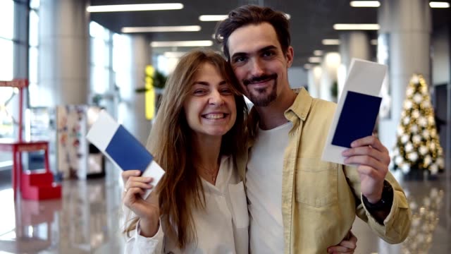 Excited-caucasian-couple-going-on-a-vacation-and-holding-passport-boarding-pass-at-airport.-Waving-with-check-in-passports,-smilling-to-the-camera,-embracing