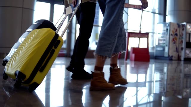 Pair-of-travelers-are-carrying-their-luggage-and-moving-over-lounge-of-terminal,-back-view.-People-are-preparing-to-boarding-and-departing.-Close-up-of-rolling-suitcases