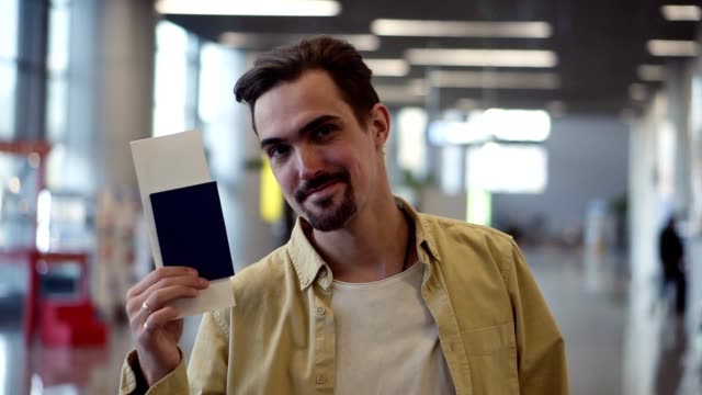 Feliz,-guapo-hombre-americano-barbudo-mostrando-pasaporte-con-billetes-de-avión-y-agitando.-Mirando-y-saqueando-a-la-cámara-mientras-está-de-pie-en-el-aeropuerto