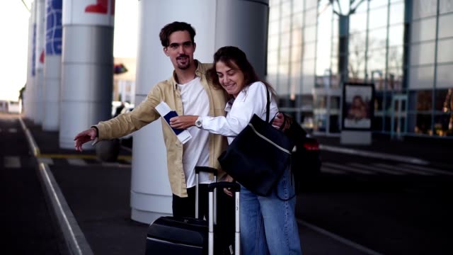 Stylish,-young-couple-with-luggage-standing-on-airport-parking-with-their-suitcases.-Waiting-for-car-after-arrival.-Man-trying-to-catch-taxi,-loving-couple-standing-embracing