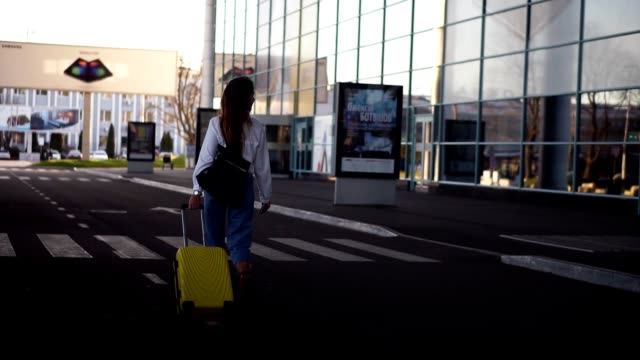 Stylish-woman-walk-with-trolley-yellow-case-by-empty-airport-terminal-outside-road,-slender-female-wearing-jeans-and-brown-boots.-Back-side-view