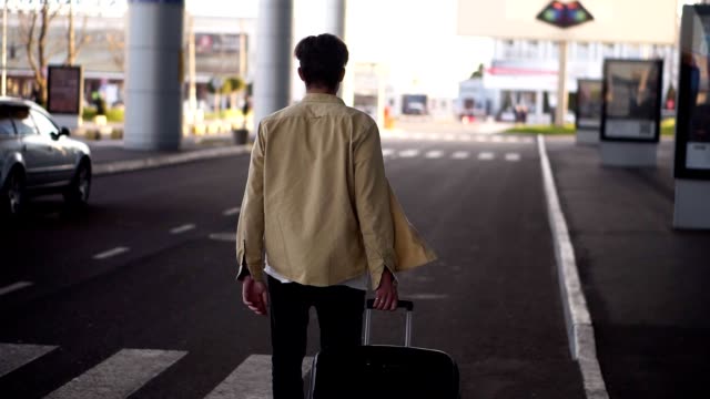 Confident,-tall-man-in-casual-with-the-black-baggage-walking-to-the-outside-airport-terminal-by-road.-Slow-motion.-Rare-view.-Blurred-backround