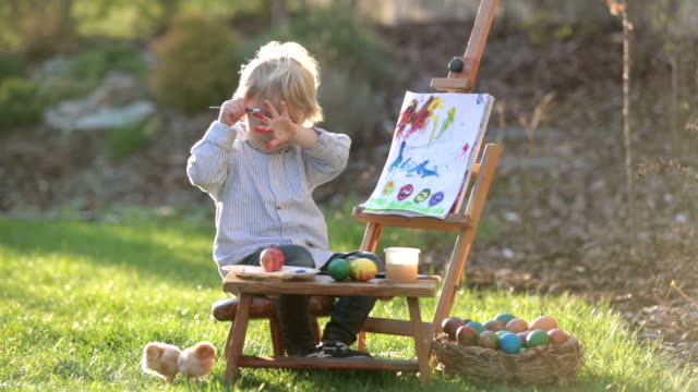 Niño-dulce,-pintando-huevos-en-el-jardín-con-polluelos-corriendo-a-su-alrededor-al-atardecer