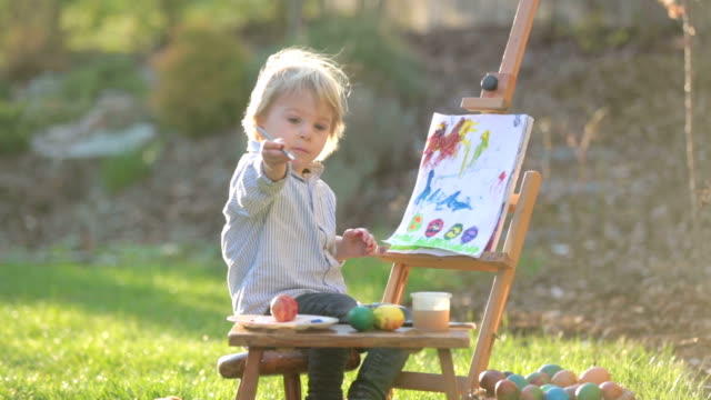 Niño-dulce,-pintando-huevos-en-el-jardín-con-polluelos-corriendo-a-su-alrededor-al-atardecer