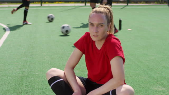 Jugadora-de-fútbol-femenina-sentada-en-el-campo-de-juego-y-posando-para-la-cámara