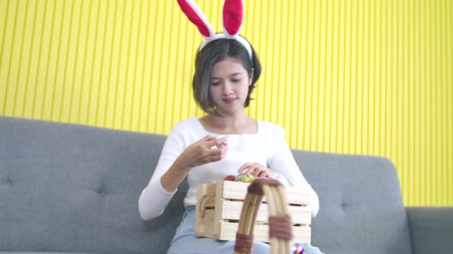 Easter,-holidays-and-people-concept.-Portrait-of-cheerful-happy-girl-wearing-bunny-ears-headband-sitting-on-the-sofa,-choosing-easter-eggs-decorated-colourful--of-handmade-in-wooden-crate-for-festival