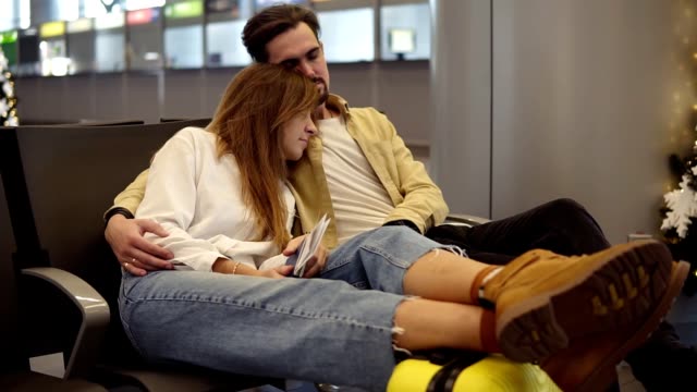 Müde-junge-kaukasische-Paar-sitzen-und-versuchen,-in-der-Abflug-Lounge-mit-Koffer-schlafen-und-warten-auf-verspäteten-Flug.-Gestresste-und-erschöpfte-Touristen,-die-auf-ihren-Flug-warten,-umarmt