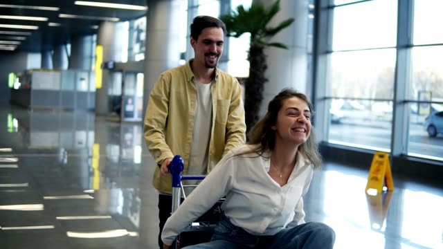 Romantic-couple-in-airport.-Attractive-young-woman-and-handsome-man-with-suitcases-are-ready-for-traveling.-Having-fun-on-luggage-trolley-while-waiting-for-departure.-Woman-immitating-flying,-smilling.-Slow-motion